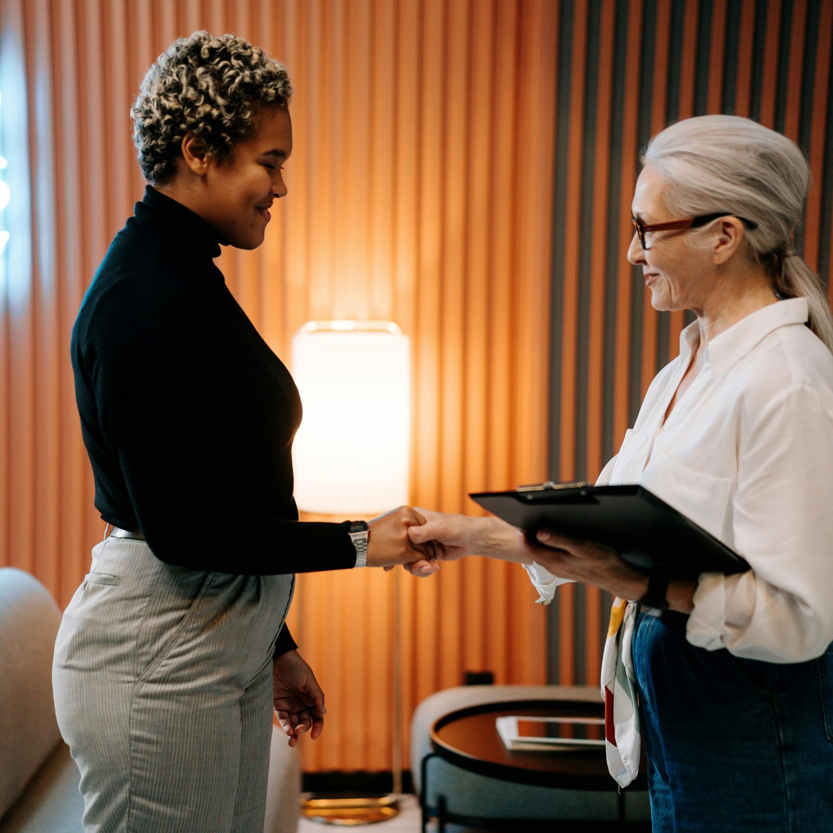 Two diverse businesswomen in a stylish office shaking hands under a 'Good Vibes Only' sign.