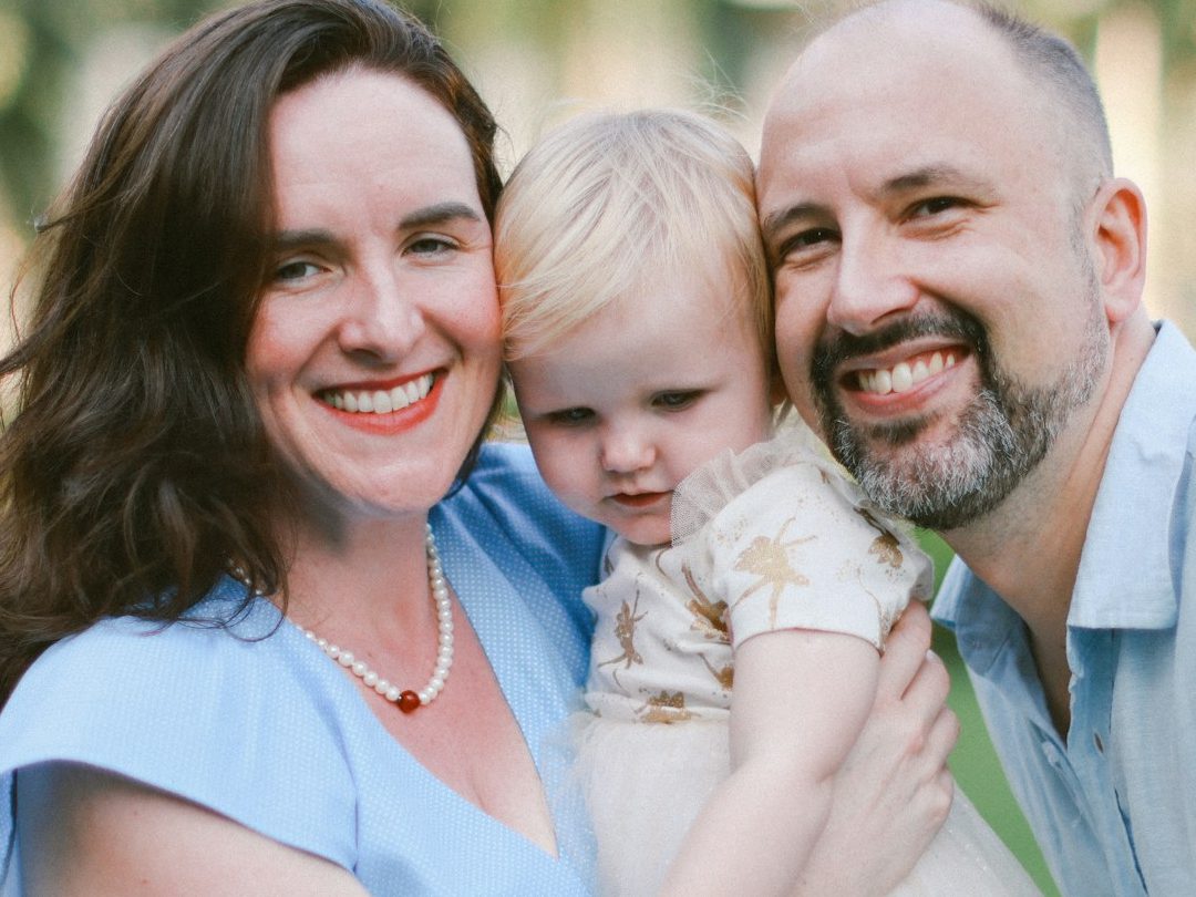 Private Household, Childcare and Education Services A family portrait featuring a woman, a man, and a toddler outdoors, smiling together.